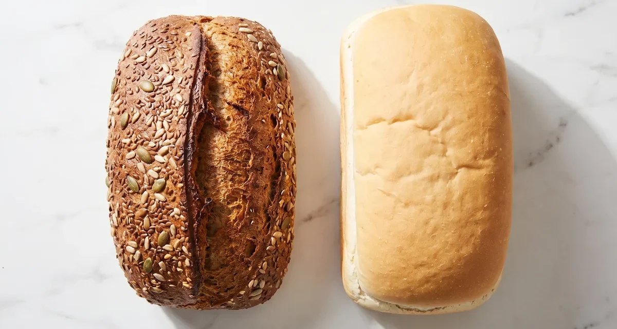 A seeded whole-grain loaf beside a pale refined white loaf, showing the fiber difference